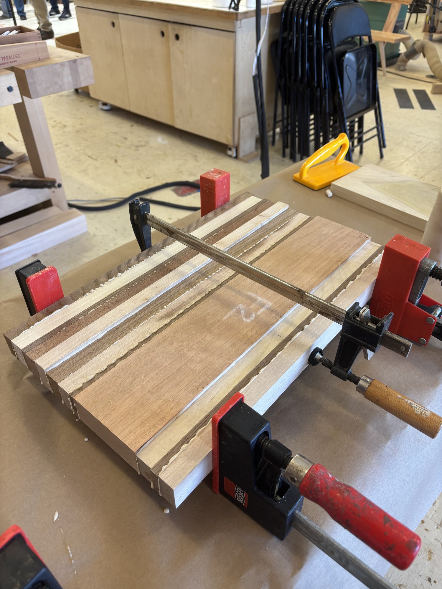 The panel in the clamps — parallel jaws front and back, with a thinner clamp laid across the top to keep the panel from bowing under side pressure. Squeeze-out is visible along every seam, which is what you want.