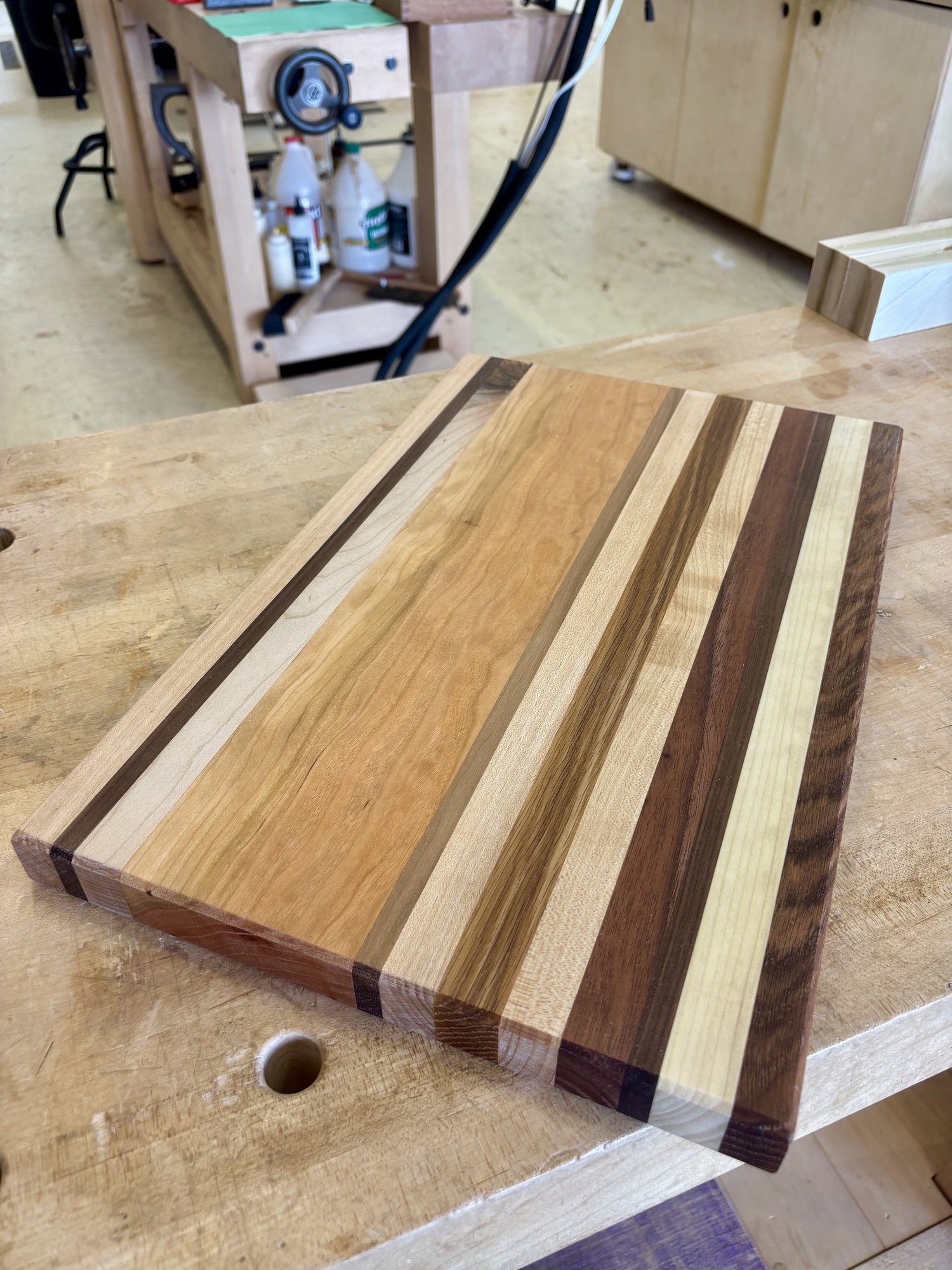 The finished cutting board on the workbench — strips of walnut, cherry, maple, and oak laminated together, oiled to a soft glow.