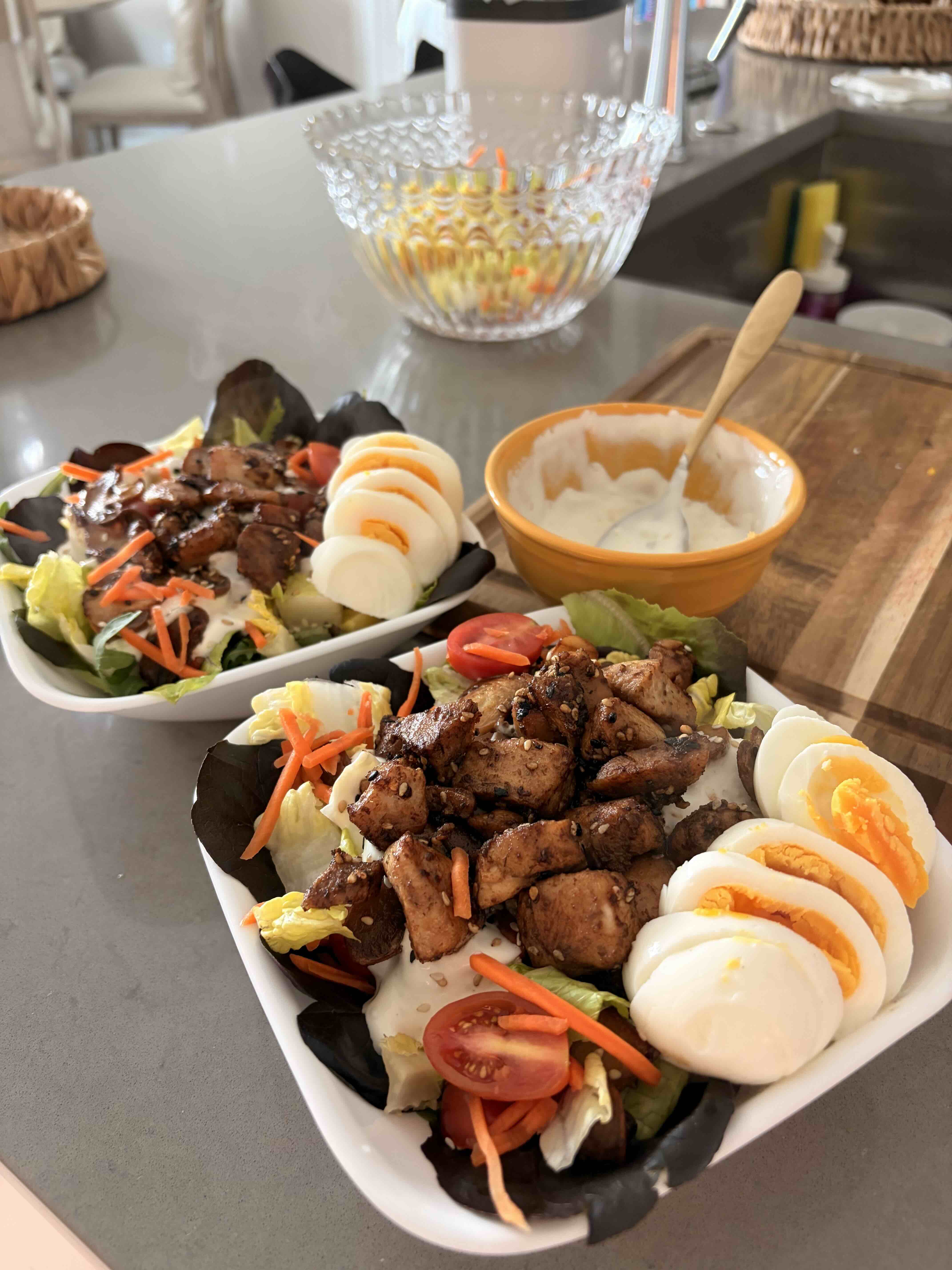 Two bowls of chicken salad with egg, tomato, and a side of dressing on a wooden board.